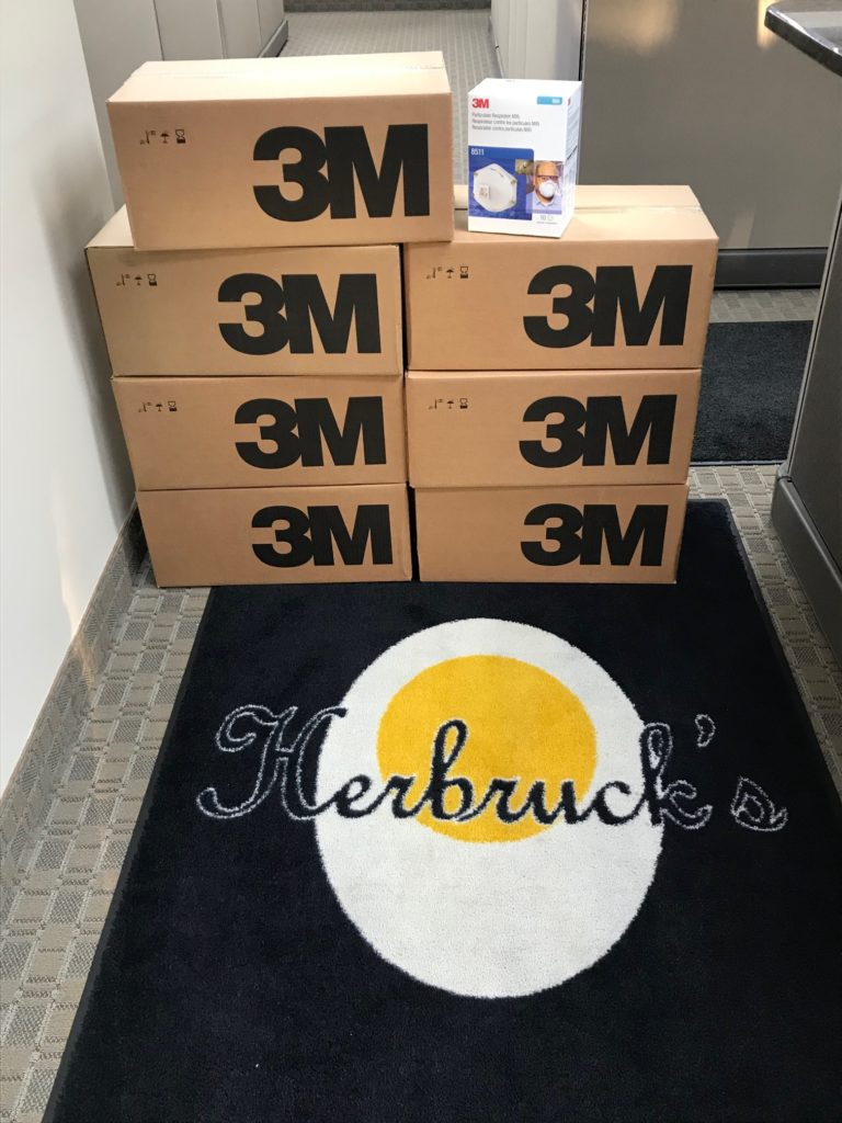 a pile of boxes with the "3M" logo on them sitting on top of a rug with the Herbruck's logo, which is an egg